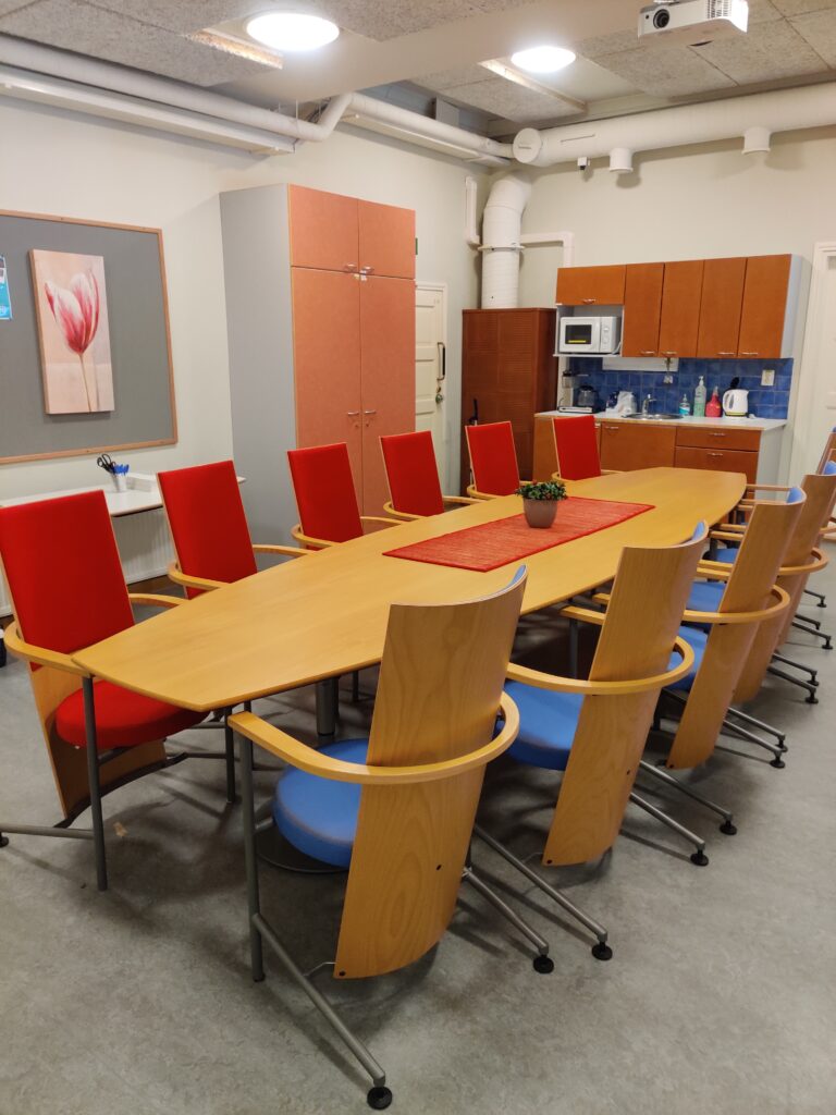 Kokoushuoneessa on pitkä pöytä, jonka toisella puolella on kuusi sinistä ja toisella puolella kuusi punaista tuolia. Valkokangas on pöydän päädyssä seinällä.