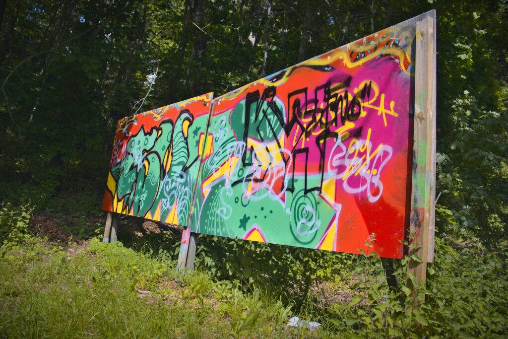 Metsän siimeksessä pylväiden varassa seisova kaksilevyinen graffitiseinä, jossa graffiteja.